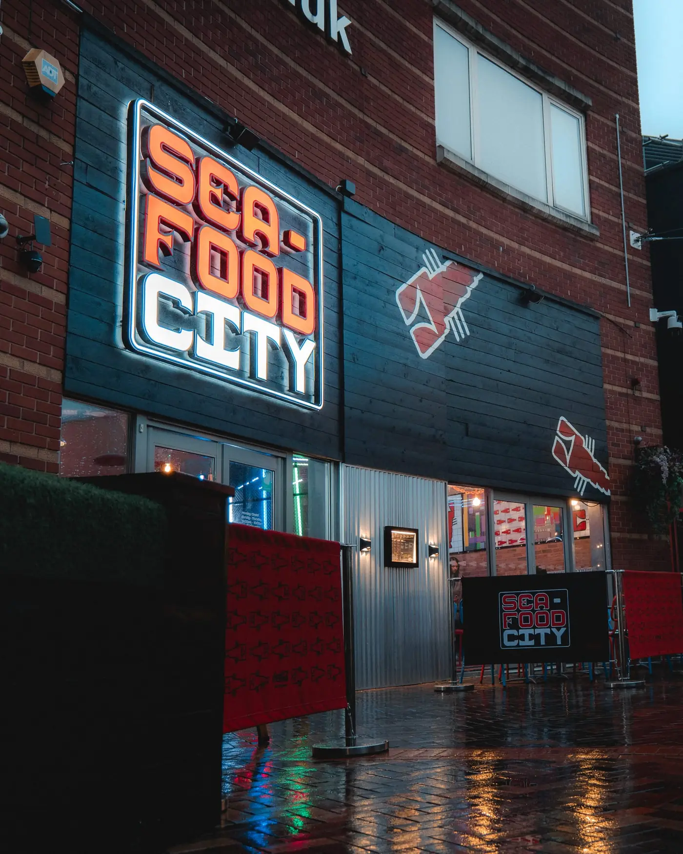 Seafood City Birmingham storefront at the Arcadian Centre at night — illuminated neon Sea-Food City sign in red, white and black, lobster decals, brick exterior, wet pavement reflecting the lights